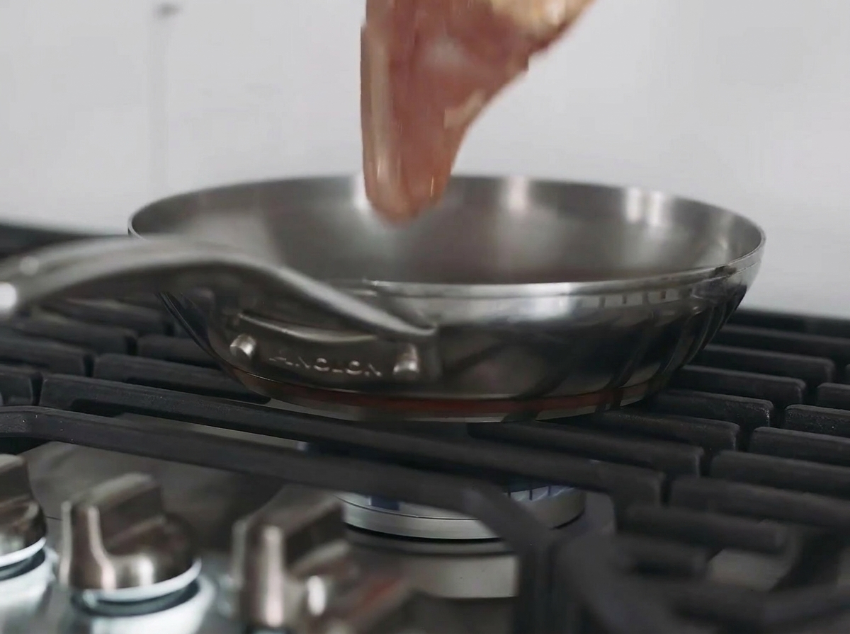 A chicken breast searing in a stainless steel pan on a stovetop.