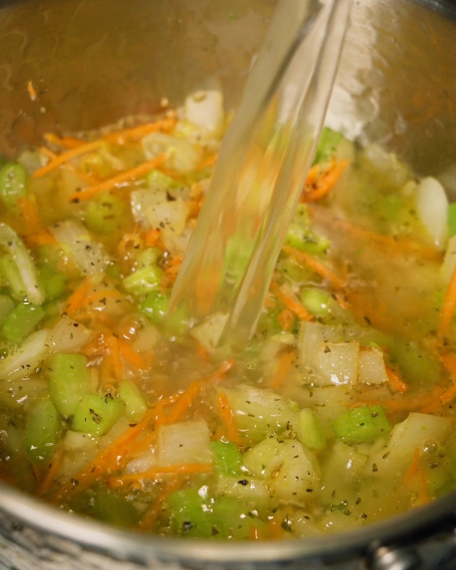 Chicken broth being poured into a pot containing sautéed vegetables and herbs.