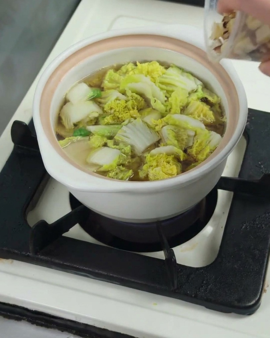 Raw chicken thigh pieces being added to a pot containing softened baby bok choy in broth.