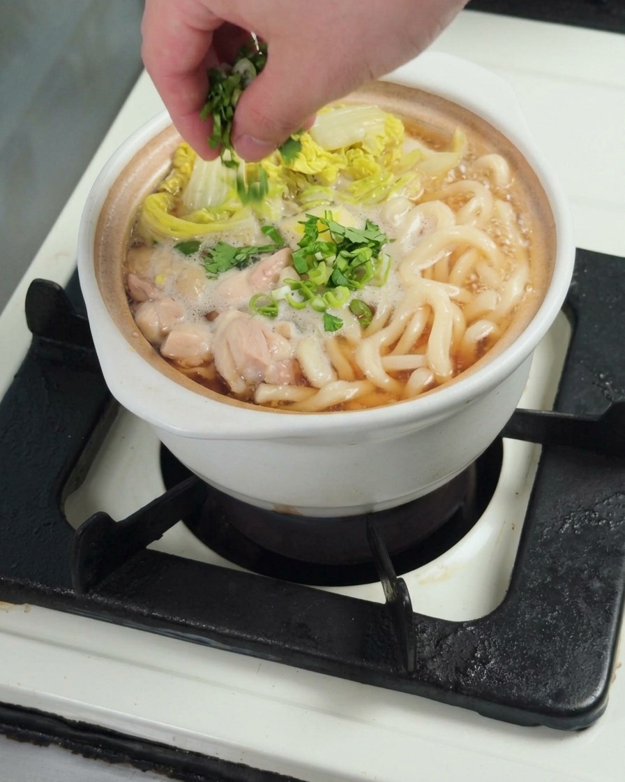 Fresh chopped green onions being sprinkled as a garnish over a finished bowl of chicken udon.