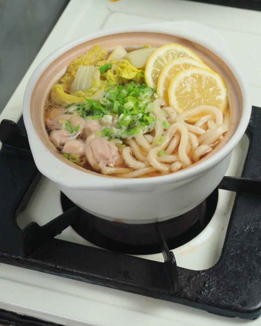 Hand squeezing a fresh lemon wedge into a pot containing cooked udon noodles and bok choy.