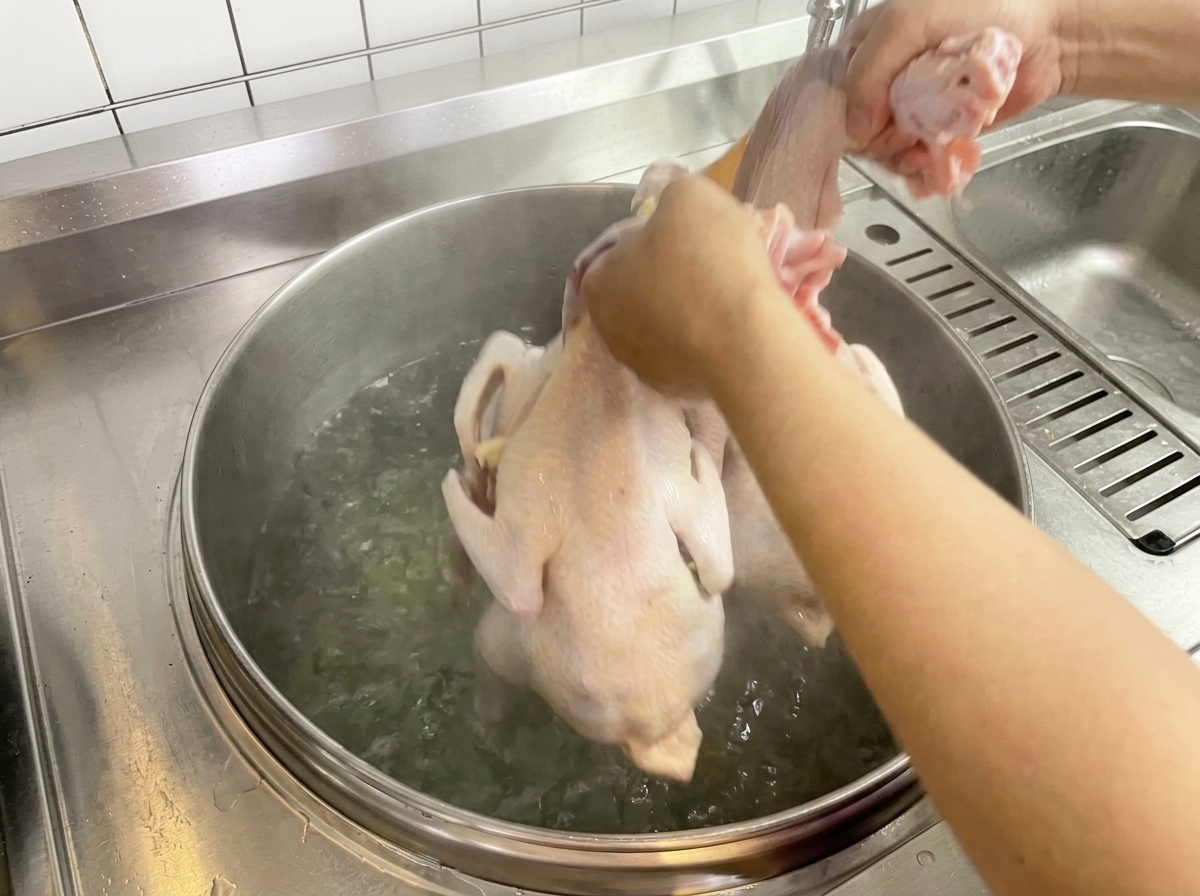 A raw chicken being dipped into a large pot of boiling water by hand.