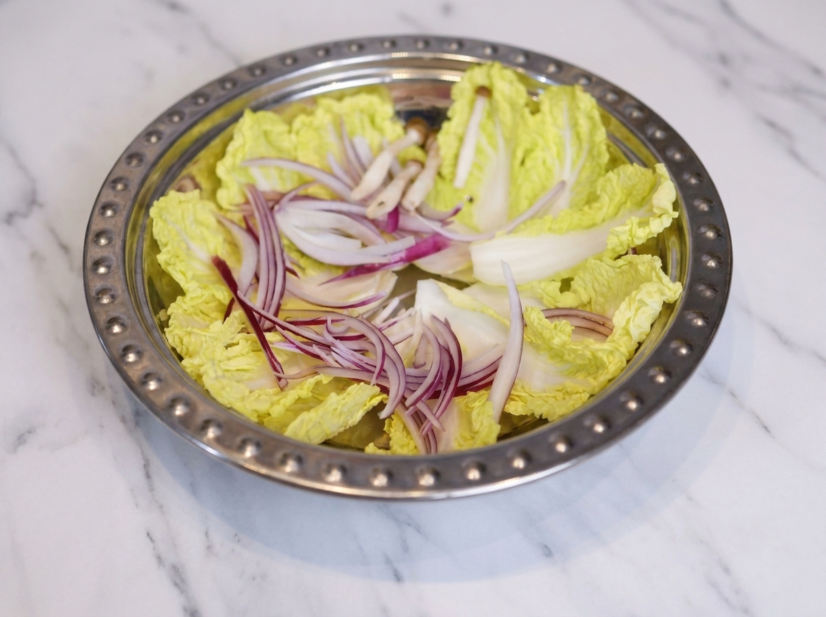 A wide metal pot filled with an organized arrangement of cabbage, onion strips, mushrooms, fish cakes, and slices of luncheon meat.
