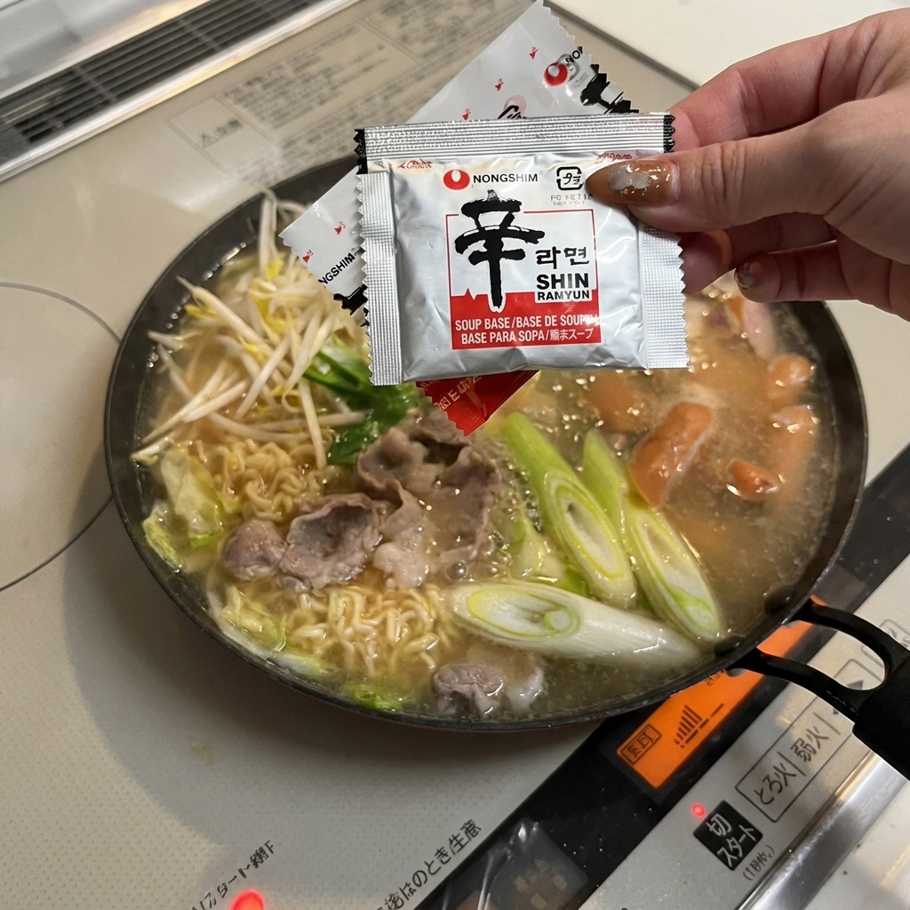 A hand emptying the red instant noodle seasoning packet into the simmering pan of stew ingredients.