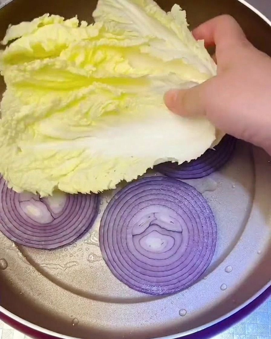 Fresh Napa cabbage leaves being placed into the pan over a layer of sautéed purple onions.