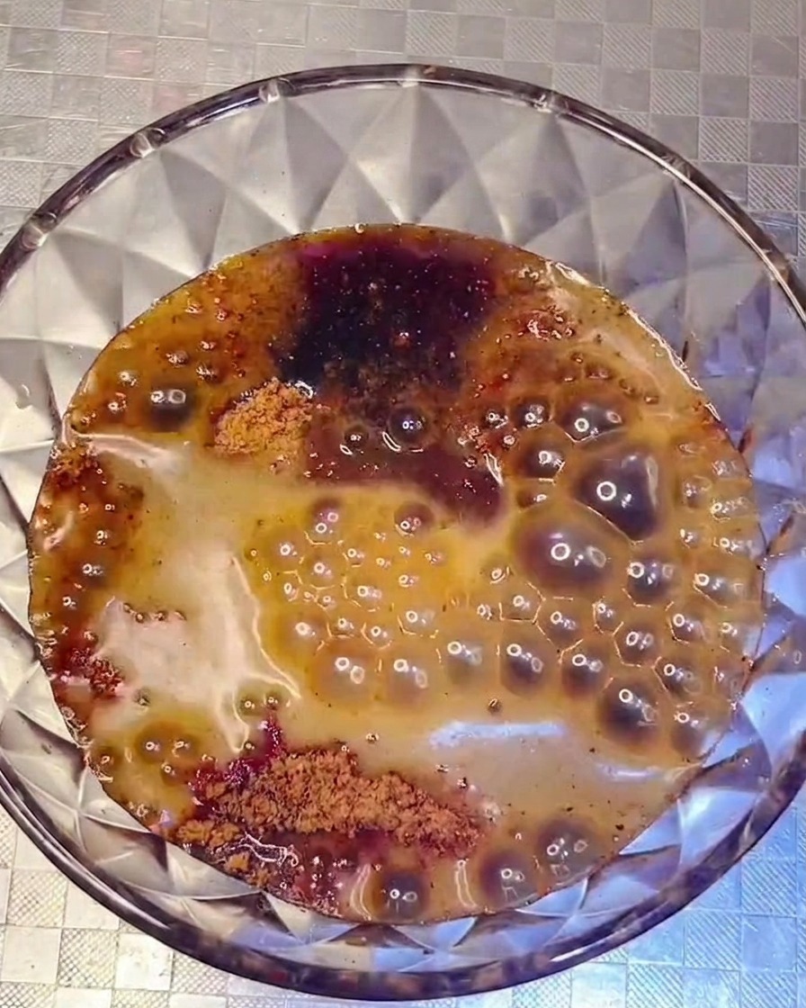 A dark, savory sauce mixture being poured from a glass bowl into a pot filled with various arranged ingredients.