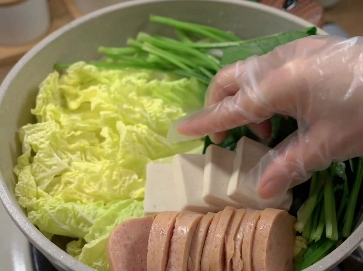 A wide pot filled with beautifully arranged layers of luncheon meat, tofu, fish cakes, and vegetables.