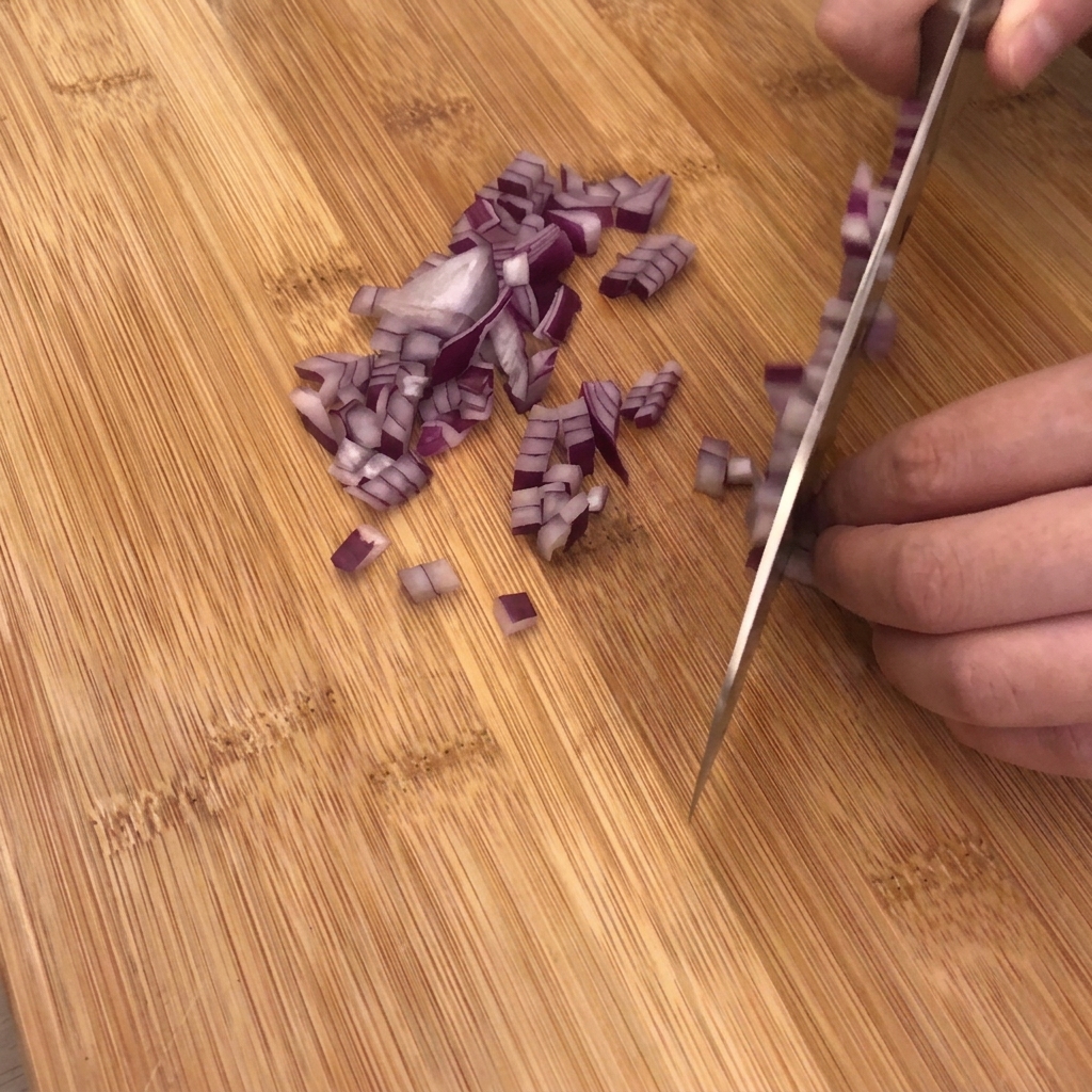 A sharp kitchen knife dicing a red onion on a wooden cutting board with garlic and bacon pieces nearby.