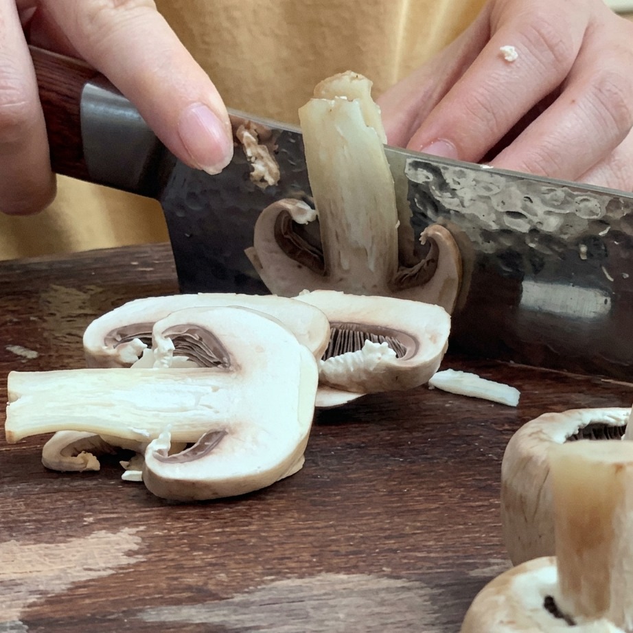 Fresh mushrooms being sliced into thin pieces on a wooden cutting board.