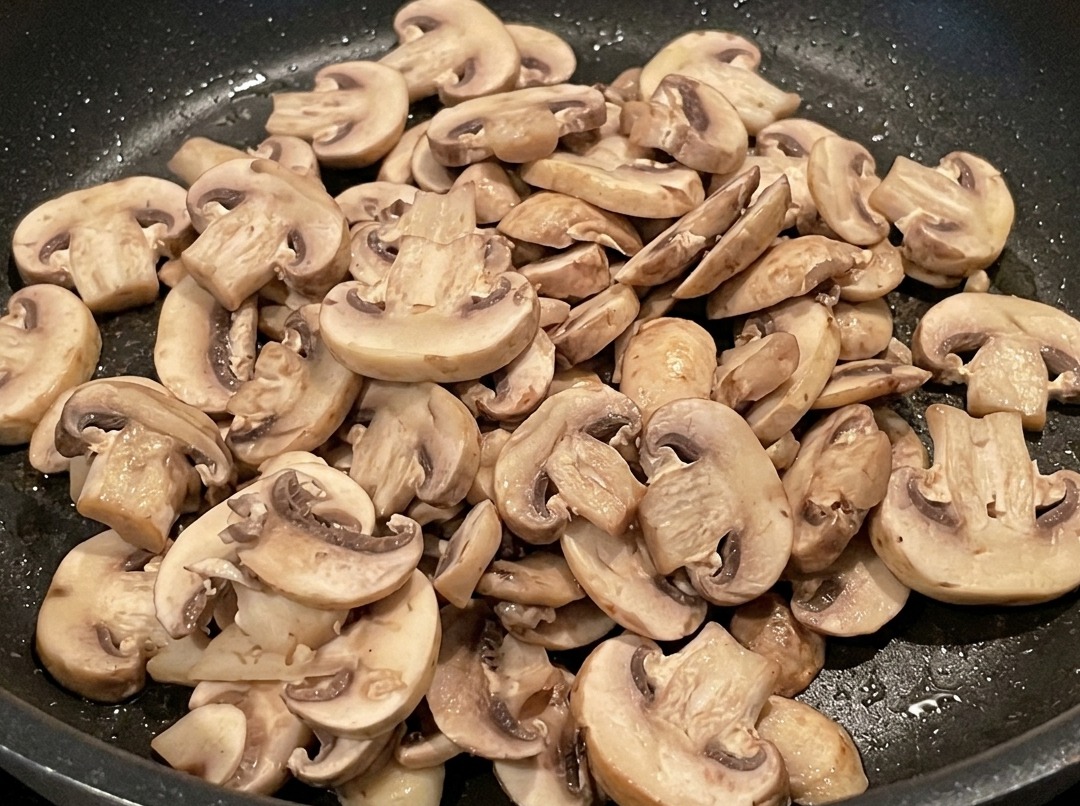 Sliced mushrooms cooking in the pan alongside rendered bacon pieces.