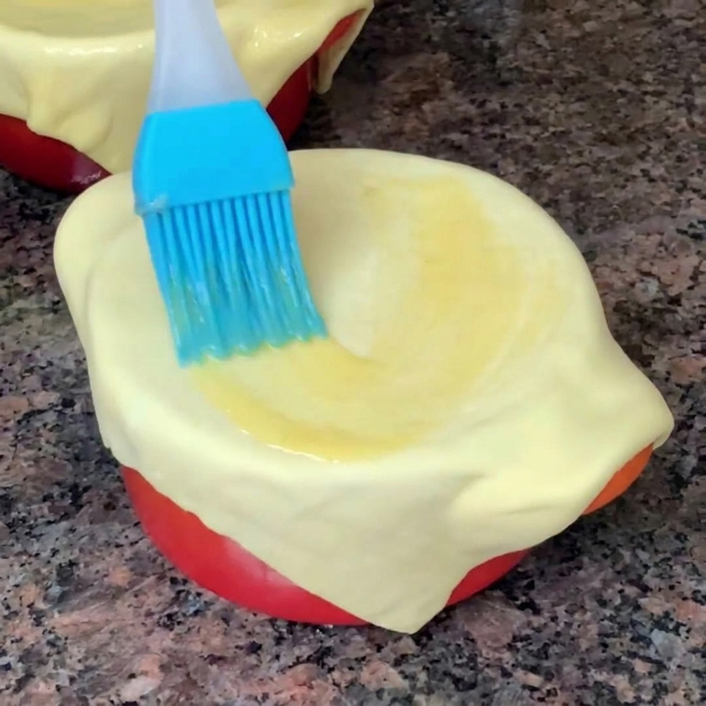 A close-up of a blue silicone brush spreading golden egg wash evenly over a raw puff pastry sheet covering a bowl.