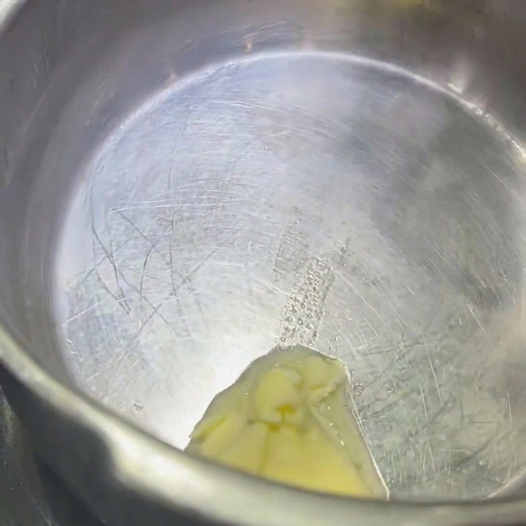 A close-up shot of a knob of butter melting in a stainless steel pot.
