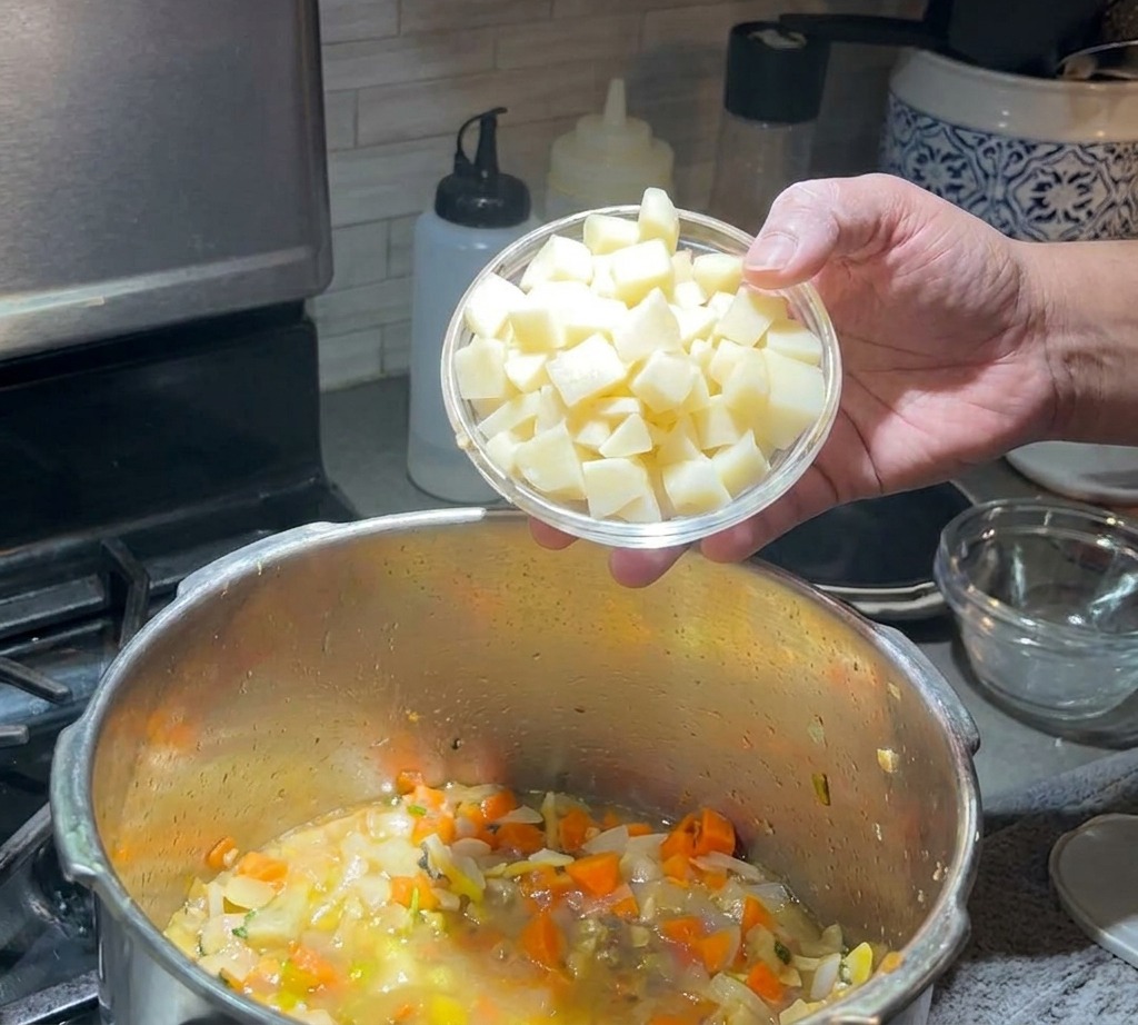 A hand pouring a small bowl of cubed potatoes into a simmering soup pot filled with carrots and onions.