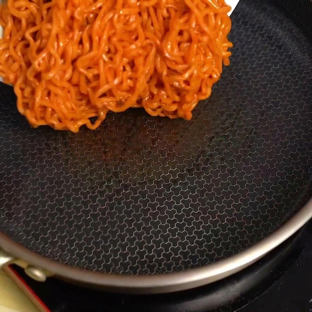 Spicy fire noodles being poured from a bowl into a hot, empty black frying pan.