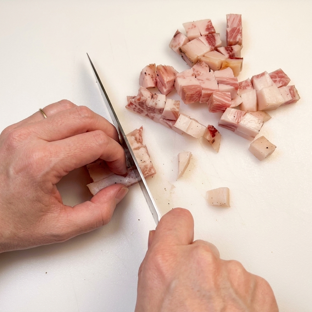 Hand with a chefs knife cutting a piece of cured bacon into small, even cubes on a white cutting board.