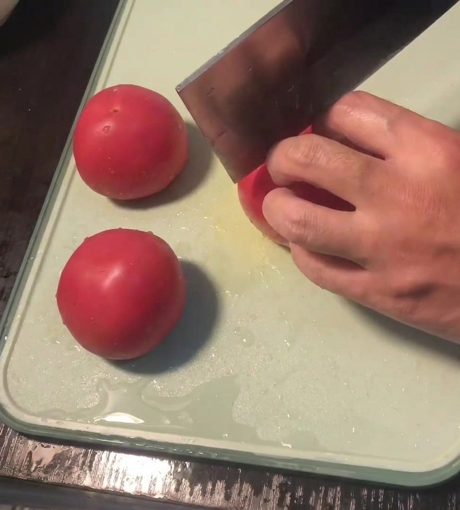 A close-up view of a hand using a knife to score a cross into the skin of a fresh tomato on a cutting board.
