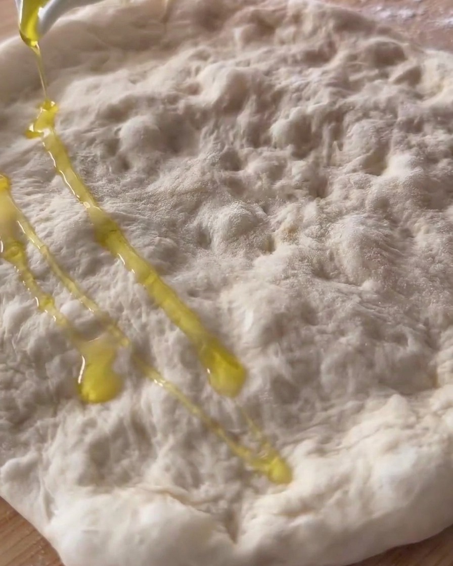 Olive oil being poured over a stretched and dimpled raw pizza dough.