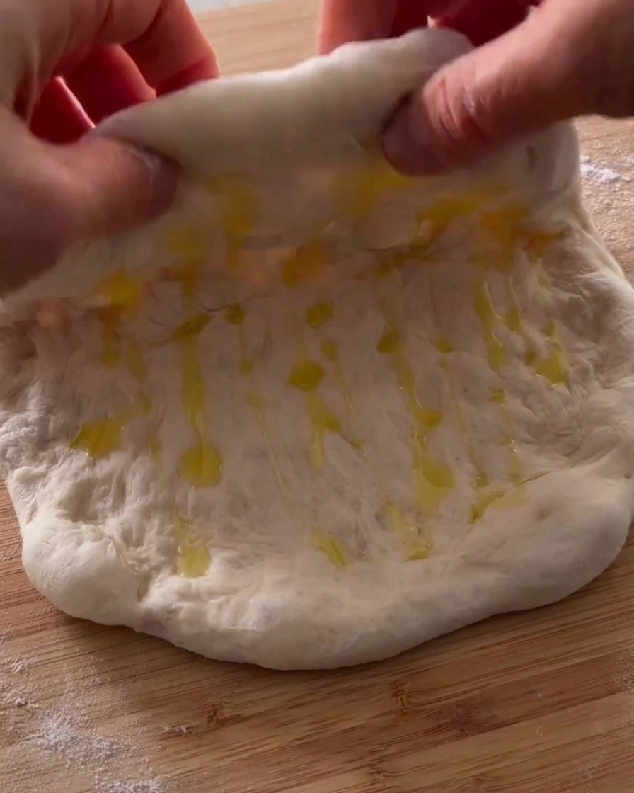 Hands folding a piece of oiled pizza dough in half on a wooden surface.