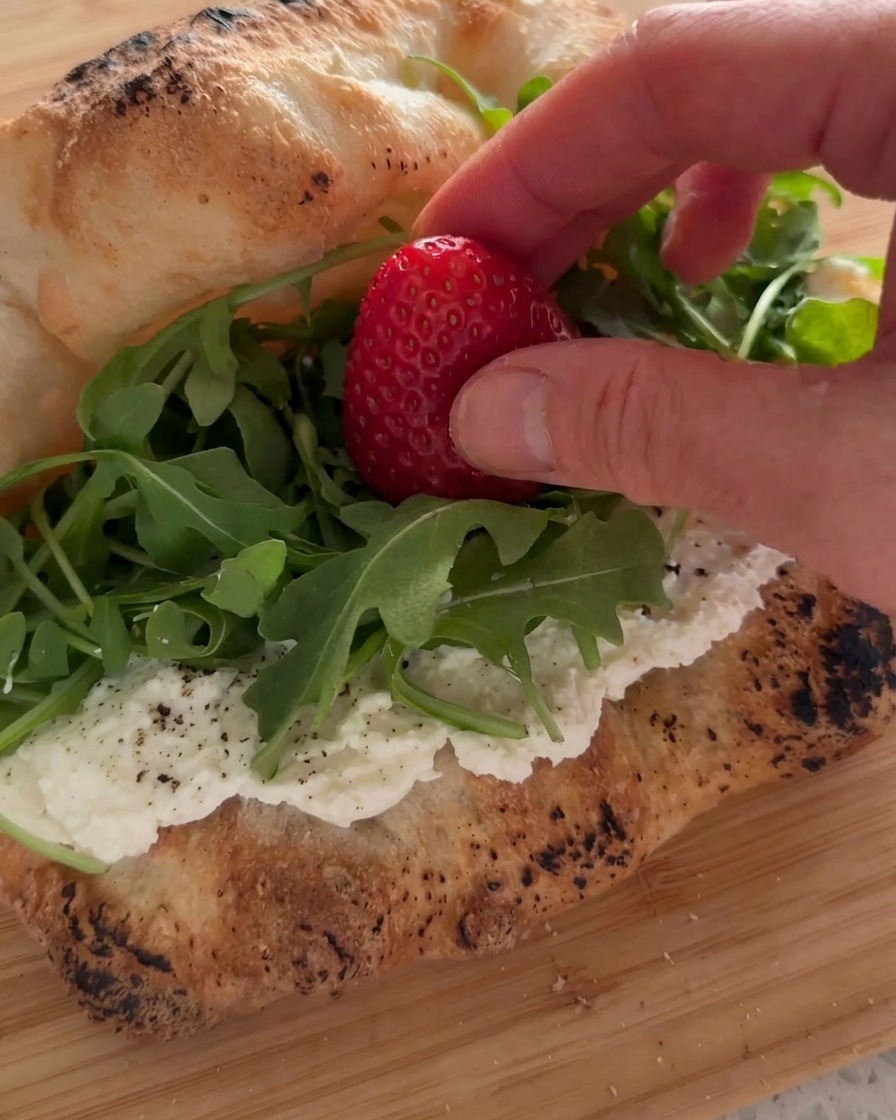 A hand placing a fresh strawberry onto a bed of green arugula inside a ricotta-lined sandwich.
