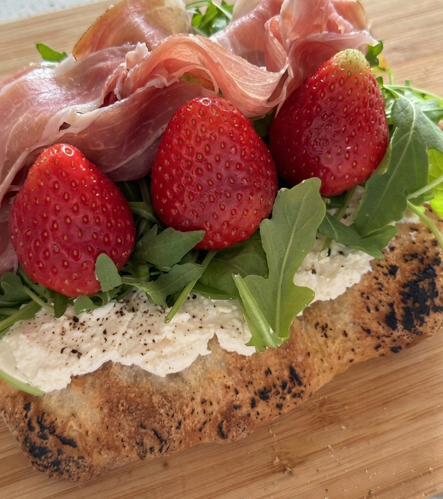 A close up of folded prosciutto slices being layered over fresh strawberries and arugula.