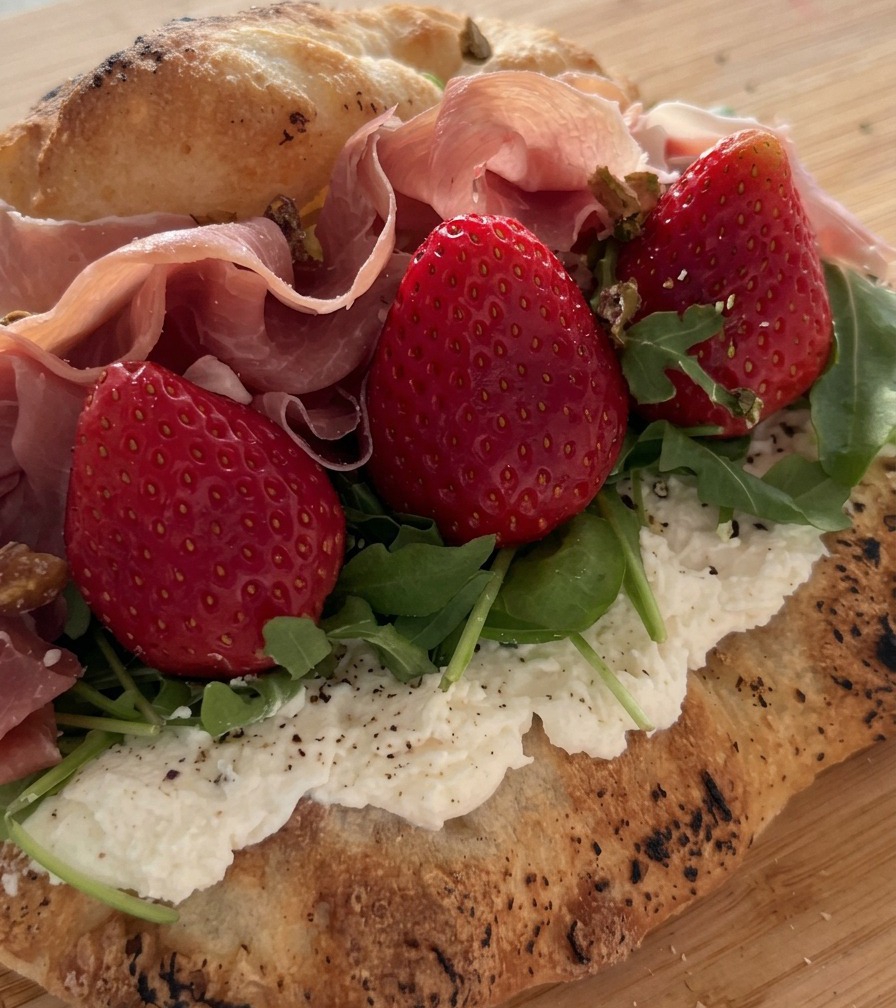 Crushed pistachios being sprinkled over a sandwich filled with prosciutto, strawberries, and arugula.
