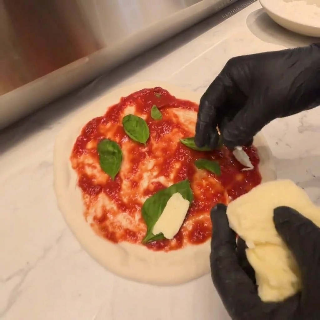 Fresh pieces of mozzarella cheese being laid over a layer of arugula on raw pizza dough.