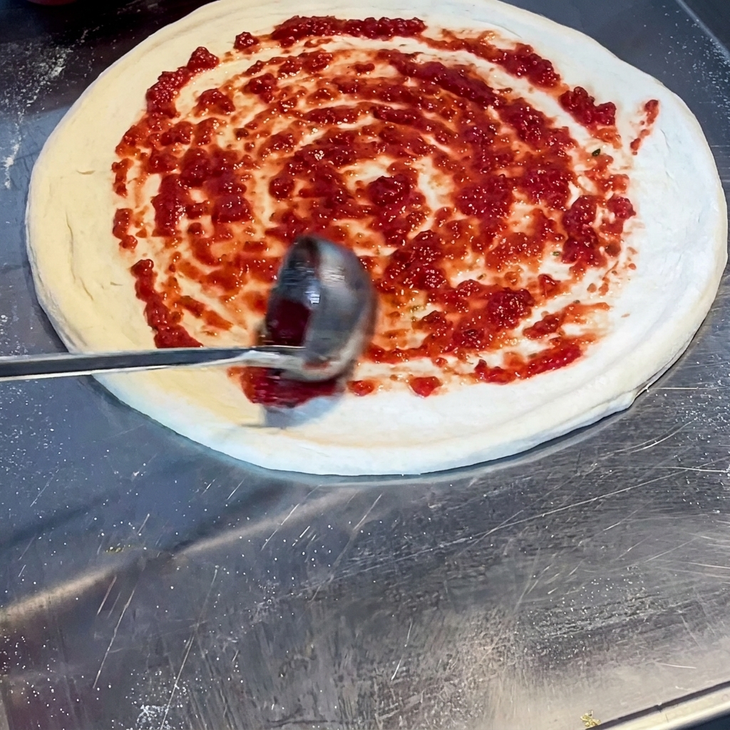 A ladle spreading vibrant red tomato sauce in a spiral pattern across the raw pizza dough.
