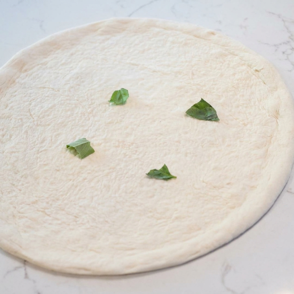 A raw, stretched pizza dough base being topped with scattered fresh green basil leaves.