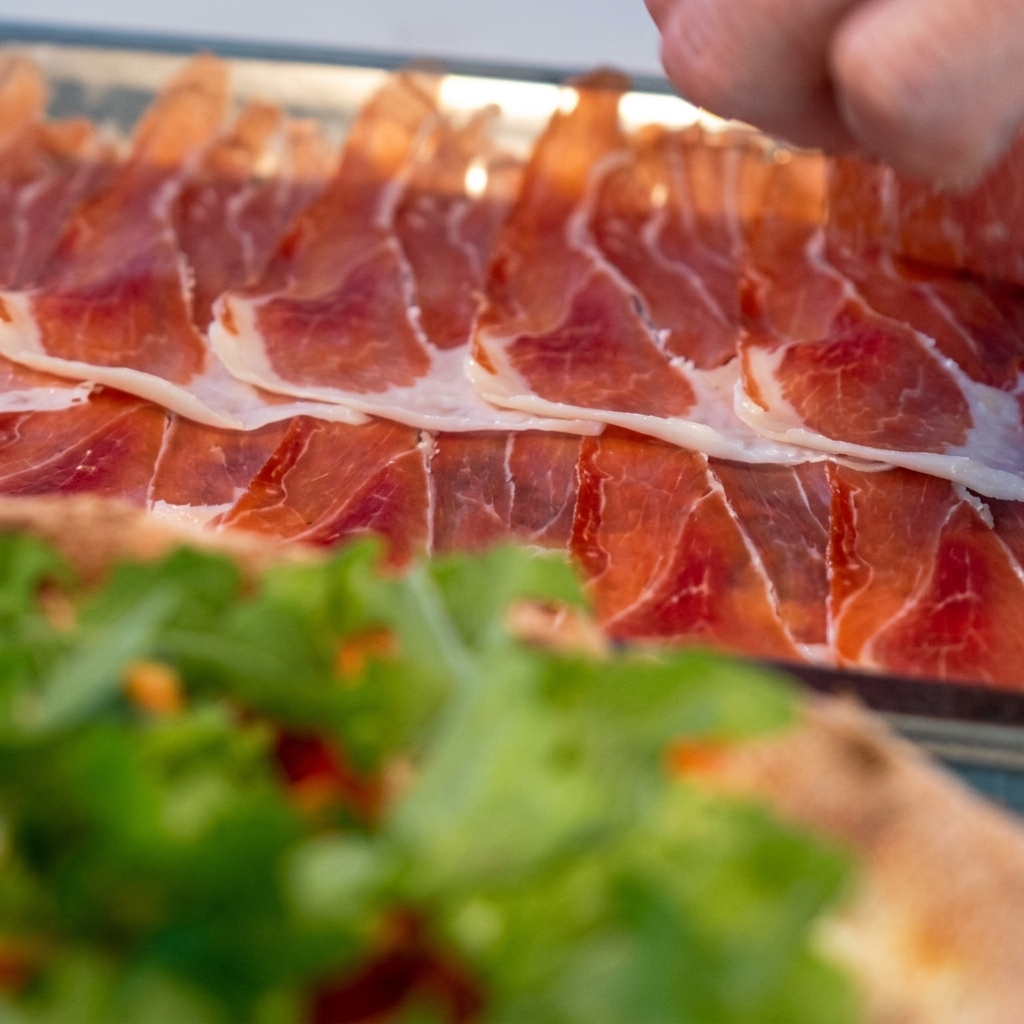 Thin slices of prosciutto being carefully arranged on top of the arugula-covered pizza.