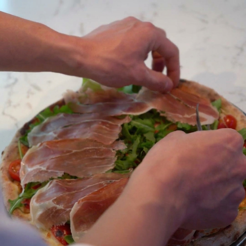 Grated parmesan cheese being sprinkled over the pizza, coating the prosciutto and arugula.