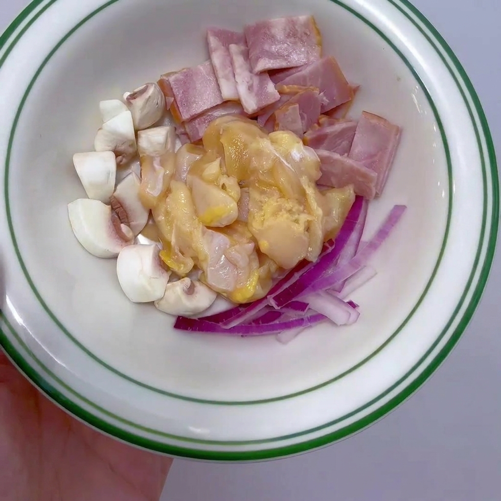 A small white bowl filled with raw diced chicken breast, sliced bacon strips, quartered white mushrooms, and thin strips of red onion.