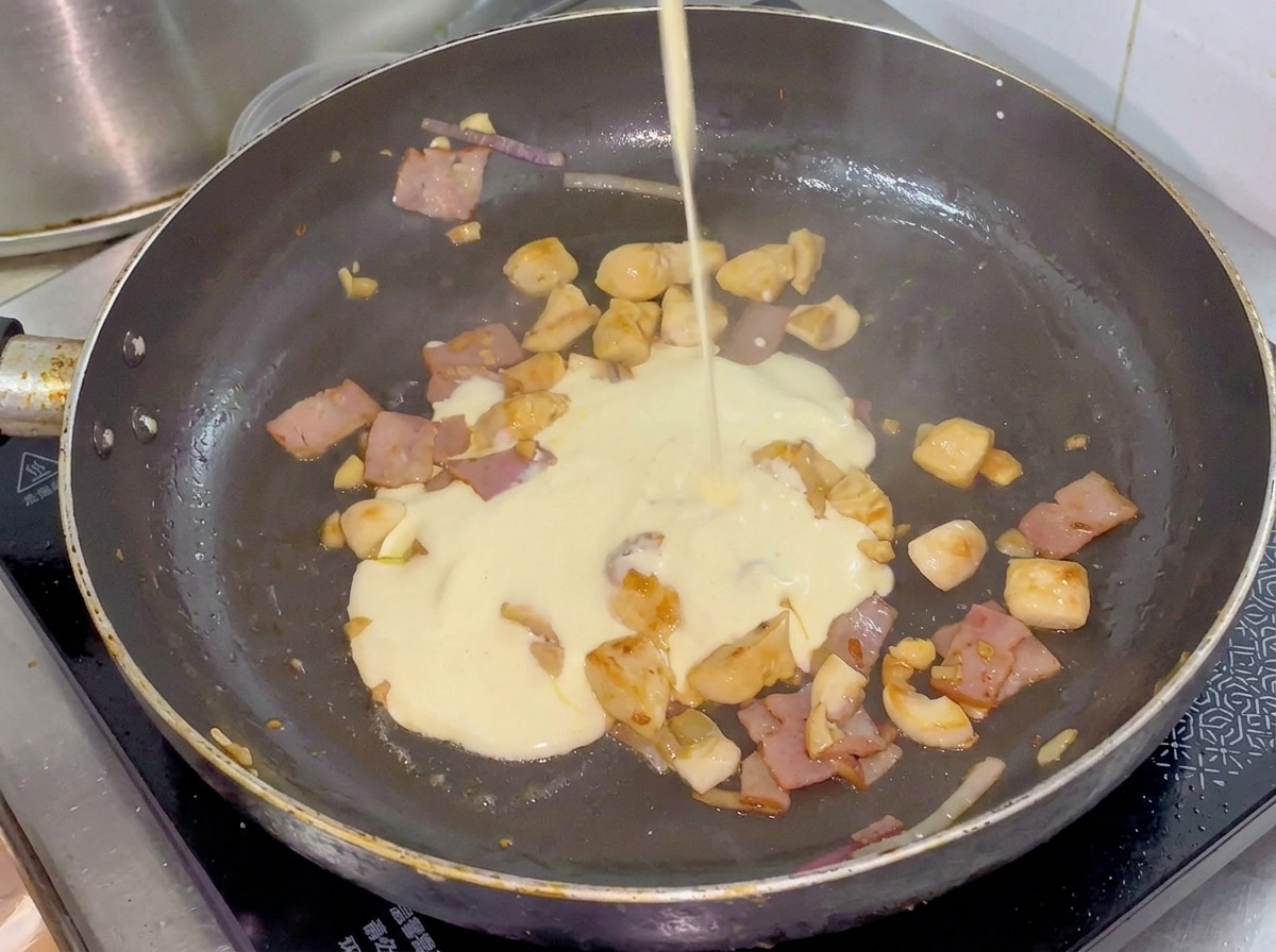 Cream being poured from a small pitcher into a pan containing cooked chicken, bacon, and vegetables.