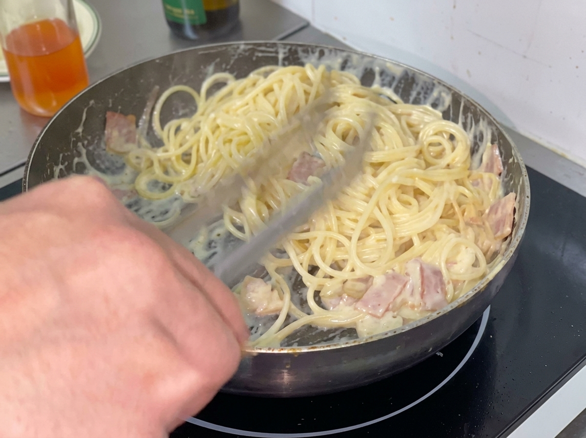 A portion of cooked spaghetti noodles being placed into a creamy sauce in a frying pan.