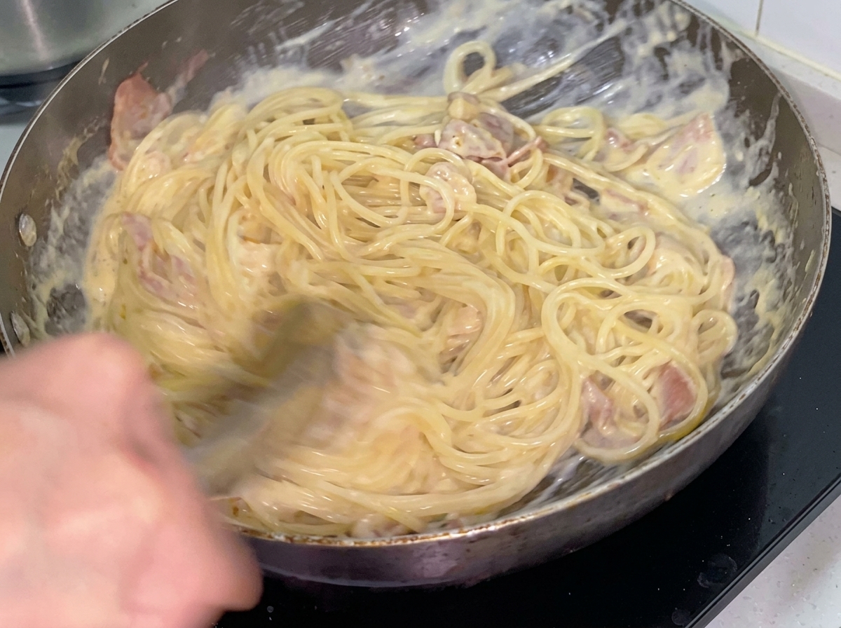 Creamy spaghetti being tossed with bacon and chicken using metal tongs in a frying pan.