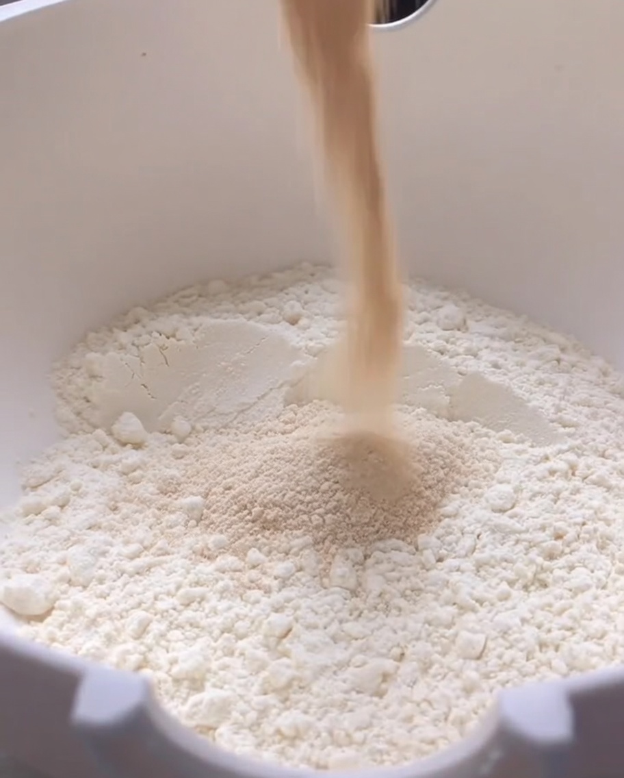 Pouring dry yeast into a large white bowl filled with flour.