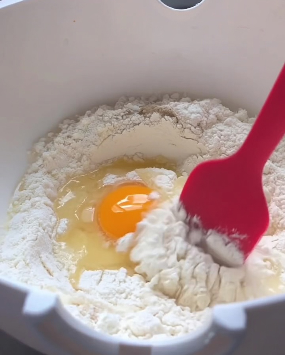 A red silicone spatula stirring dry white flour, a cracked raw egg, and warm water together in a white mixing bowl.