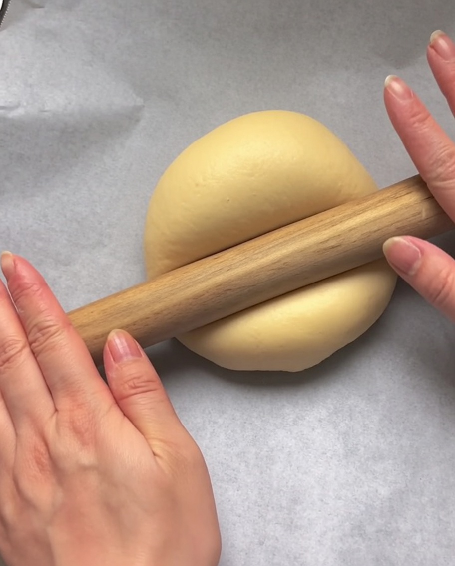 Hands using a wooden rolling pin to flatten a ball of raw dough into a circular flatbread shape on white parchment paper.