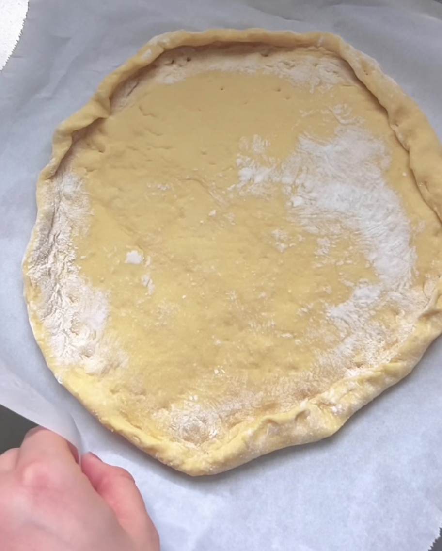 A hand folding and pinching the edge of the flattened raw dough to form a raised crust rim on parchment paper.