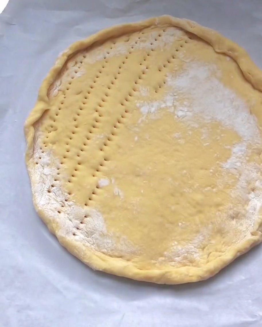 A raw circular pizza crust on parchment paper with small holes poked evenly across its flat center surface.