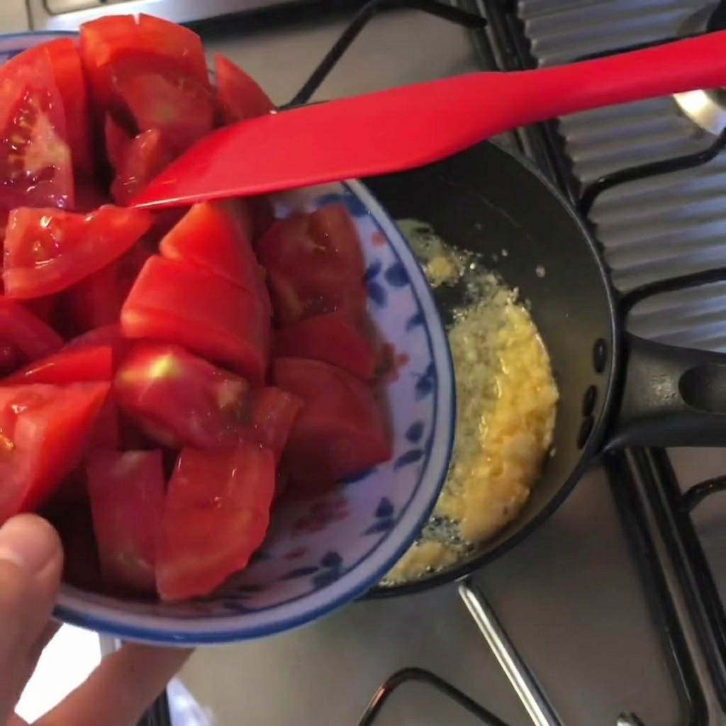 Fresh red tomato chunks being added to a black pot with melted butter and minced garlic.