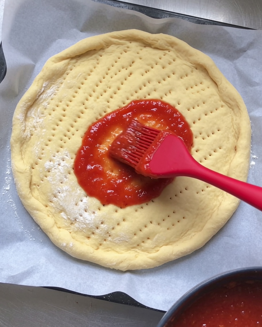 Red tomato sauce being spread evenly over a round, dimpled raw pizza dough base.