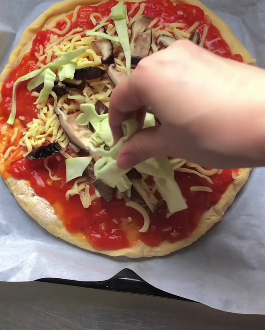 A hand placing shredded raw green cabbage over a pizza layered with tomato sauce, cheese, and sliced mushrooms.