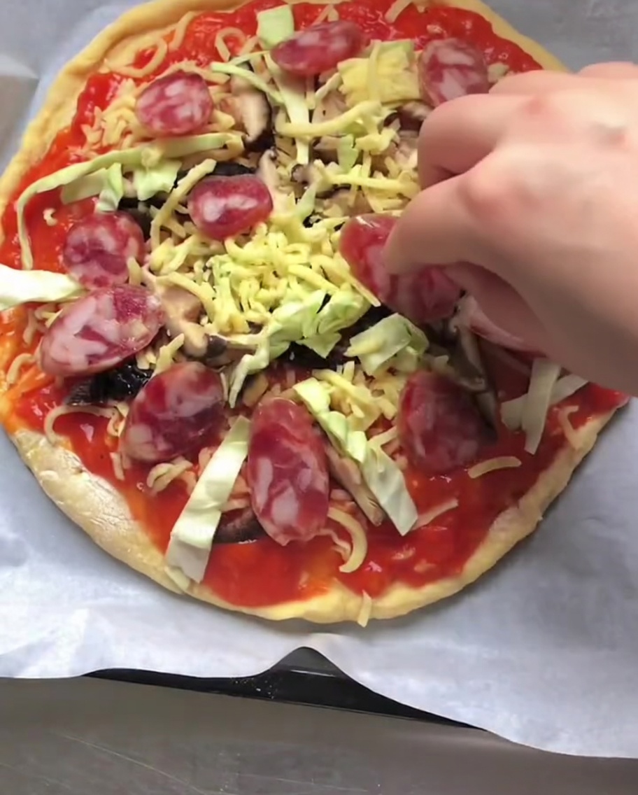 A hand placing slices of red Italian sausage onto an uncooked pizza topped with sauce, cheese, and vegetables.