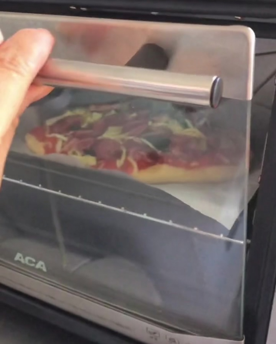 Opening the door of a small countertop oven to place a raw, assembled pizza inside on a baking tray.
