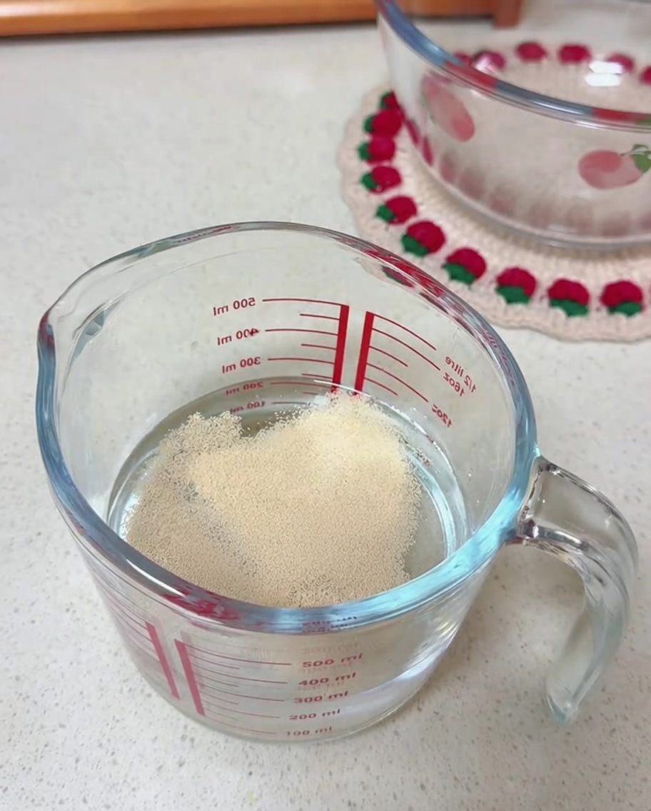 Dry yeast granules resting on the surface of water inside a clear glass measuring cup.