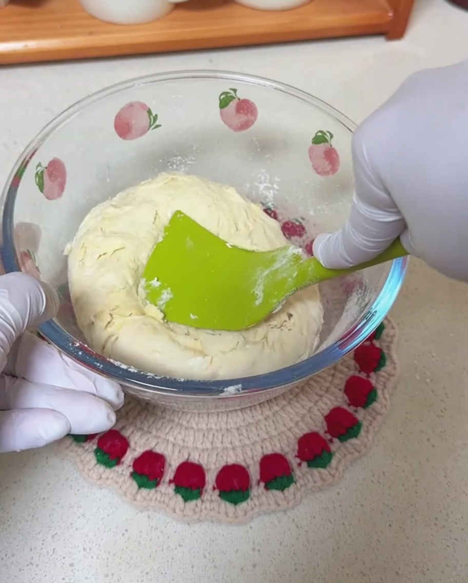 Gloved hands using a green silicone spatula to mix a rough dough inside a glass bowl.