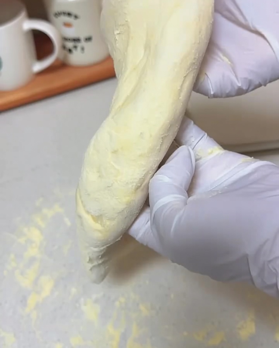 Gloved hands holding a soft piece of dough vertically, allowing it to stretch into a thin pizza crust shape.