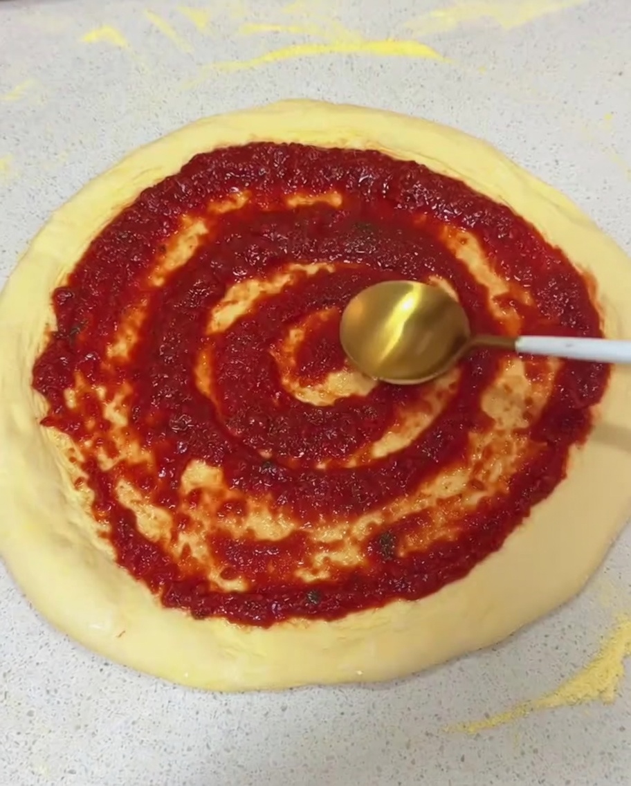 Spreading red tomato sauce in a circular motion on a stretched raw pizza dough using the back of a spoon.