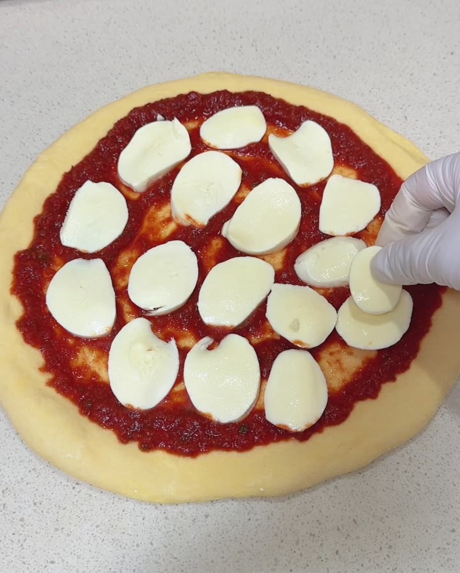 A gloved hand placing thick slices of fresh mozzarella cheese onto a pizza base covered in red tomato sauce.