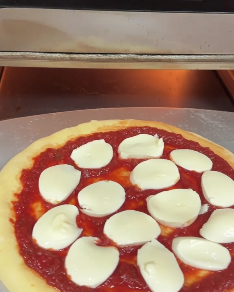 An uncooked pizza with sauce and mozzarella cheese being slid into an electric pizza oven on a metal peel.